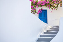 White Cycladic Architecture With Blue Door And Pink Bougainvillea Flowers On Santorini Island, Greece. Fantastic Travel Background Idyllic Summer Vacation Holiday Concept. Wonderful Summer Luxury Vibe