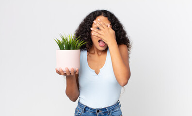 hispanic pretty woman looking shocked, scared or terrified, covering face with hand and holding a houseplant