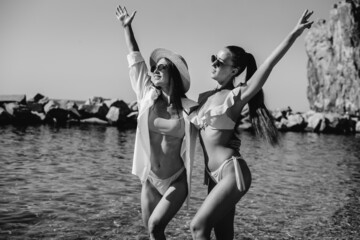 Two beautiful young girls in glasses and bikinis are standing and enjoying themselves on the ocean shore against the background of huge rocks on a sunny day. Tourism and tourist trips. Black and white