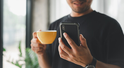 Happy Asian man using smartphone and drinking coffee he's smiling and relax in living room at home, man using cellphone for searching data and social media on internet. Technology and people concept.