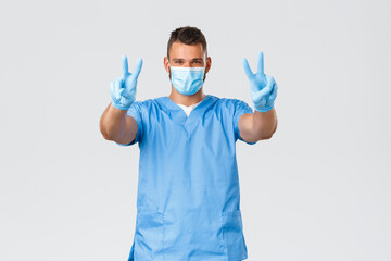 Wall Mural - Healthcare workers, covid-19, coronavirus and preventing virus concept. Handsome hispanic doctor, nurse in scrubs and medical mask with gloves, show peace signs, stay positive