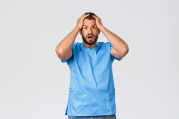 Healthcare workers, medicine, covid-19 and pandemic self-quarantine concept. Shocked and nervous male doctor, nurse in blue scrubs, gasping astonished, hold hands on head startled