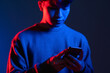 © golubovy - Modern gadget. Virtual lifestyle. Online communication. Social network. Focused Asian man internet browsing on phone in red blue color neon light shine in dark gradient studio.