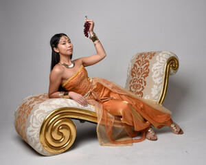 Full length portrait of pretty young asian woman wearing golden Arabian robes like a genie, seated pose on lounge, isolated on studio background.