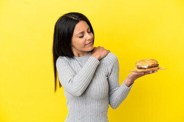 Young caucasian woman holding a burger isolated on yellow background suffering from pain in shoulder for having made an effort