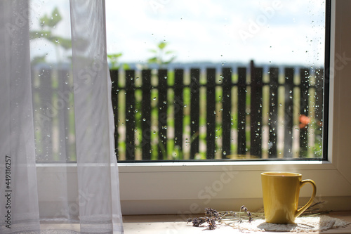 yellow mug with a hot drink on the windowsill against the background of the window after the rain. cozy summer morning in the village