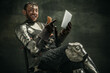 One brutal bearded man, medeival warrior or knight with digital tablet sitting on chair over dark background.