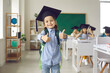 © Studio Romantic - Schoolboy is wearing a graduation cap, showing thumb up. Schoolboy stands on blurred background of class, graduation, school board, back to school. Smart elementary school kid