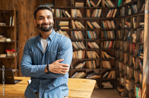 Smiling bearded indian businessman stands near desk and looks at the camera. Young positive male student in library with bookshelves on background. Proud and successful mixed-race small business owner