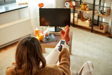 Poster - halloween, holidays and leisure concept - young womanwith remote control watching tv and resting her feet on table at cozy home