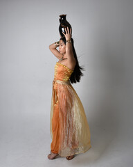 Full length portrait of pretty young asian woman wearing golden Arabian robes like a genie, standing pose holding a vase, isolated on studio background.