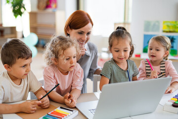 Group of small nursery school children with teacher on floor indoors in classroom, using laptop.