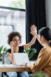 © LIGHTFIELD STUDIOS - happy interracial businesswomen giving high five while working near laptop at workplace