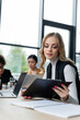 © LIGHTFIELD STUDIOS - blonde businesswoman looking at clipboard while working near laptop and blurred interracial colleagues