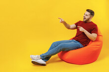 Full Length Smiling Happy Young Man In Red T-shirt Casual Clothes Sit In Bag Chair Point Index Finger Aside On Workspace Area Mock Up Isolated On Plain Yellow Color Wall Background Studio Portrait