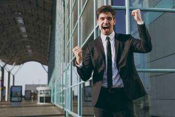 Wall Mural - Happy young traveler businessman man 20s in black classic tie suit standing outside at international airport terminal do winner gesture clench fist People air flight business trip lifestyle concept.