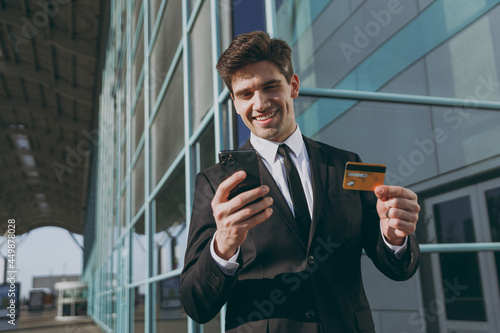 Bottom view fun young traveler businessman man in black suit stand outside at international airport terminal use mobile cell phone credit bank card do online payment Air flight business trip concept.