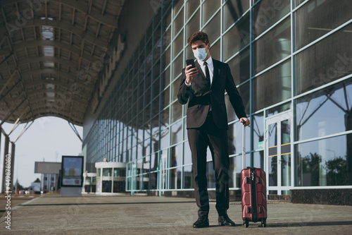 Bottom view traveler businessman young man in black suit facial mask stand outside at international airport terminal with suitcase use mobile phone book taxi order hotel Air flight business concept