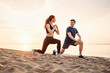 © ViDi Studio - Full size couple young two friend strong sporty sportswoman sportsman woman man in sport clothes warm up training do lunges do exercise on sand sea ocean beach outside on seaside in summer day morning
