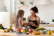 © deniskomarov - Beautiful young mother reading to adorable daughter on kitchen. Mom and child learning recipe for cooking dinner. Happy family engaged in food preparation at home. Girl sit on table full of vegetable