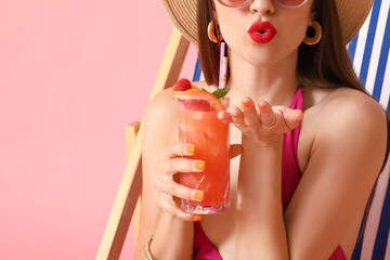 Wall Mural - Beautiful young woman in swimsuit and with cocktail blowing kiss on color background