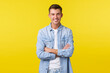 Portrait of handsome happy caucasian guy with white teeth, smiling broadly, cross arms chest confident, standing yellow background in denim shirt over white t-shirt