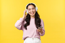 People Emotions, Lifestyle Leisure And Beauty Concept. Happy Smiling Asian Woman Listening Music In Wireless Headphones And Looking Pleased With Nice Sound, Holding Mobile Phone
