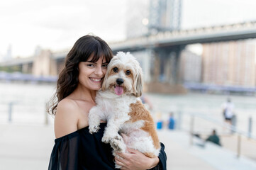 Wall Mural - a woman holding her small mixed breed dog at the boardwalk by the hudson river with brooklyn bridge and new york city buildings in the background