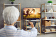 © Studio Romantic - Senior woman watching photo collection of her life memories, family, children and grandchildren, or movie, series or serial on television channel on widescreen TV at home. Back rear view from behind