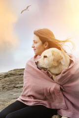 A girl with her dog is sitting on the beach covered with a blanket.