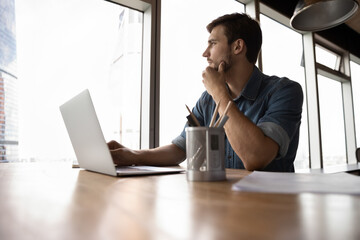 Thoughtful employee, business man, manager looking out of window in deep thought, sitting at workplace with laptop in office, thinking over company future, pondering on decision for work task