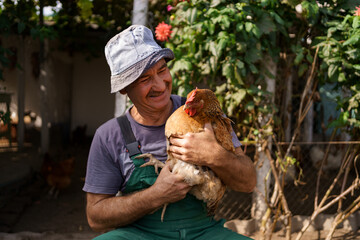 Portrait of happy caucasian farmer holding a brown hen outdoor. Smiling mature man with chicken in hand with copy space.