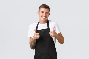 Wall Mural - Employees, grocery stores and coffee shop concept. Handsome friendly guy worker in cafe, store clerk wearing black apron, showing thumbs-up and smiling to welcome guest, guarantee quality