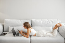 Young Woman Lying Relaxed With Phone And Laptop On The Comfortable Couch At Home. Front View. Working Or Leisure Time At Home