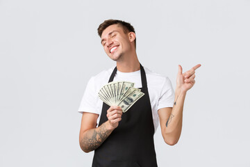 Wall Mural - Small retail business, payments and employees concept. Joyful happy part-time cafe staff, waiter in black apron close eyes and smiling as pointing right, holding money, white background