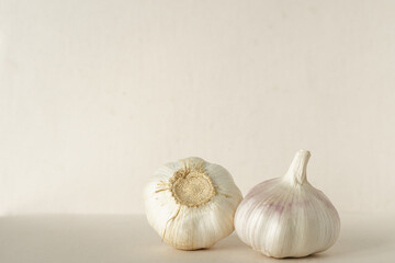 Two heads of garlic lay on a cream-colored ground against a cream-colored wall with soft light of the morning.