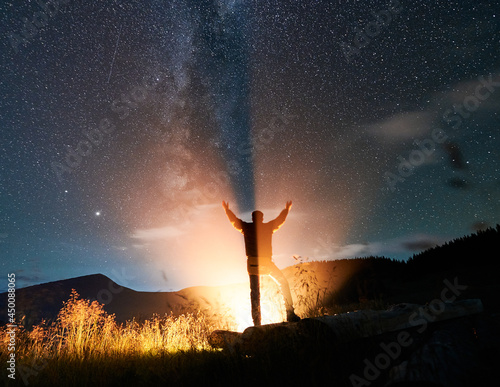 Back view of man hiker looking at night starry sky and raising hands while bonfire illuminating male silhouette. Traveler reaching out his hands to night sky with stars. Concept of travelling.