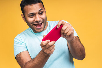 Portrait of a cheerful young african black indian man dressed in casual playing games on mobile phone isolated over yellow background.