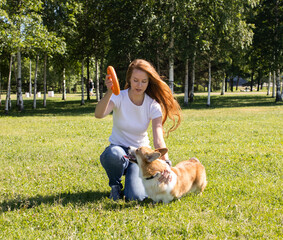 A girl with long red hair, in a white T-shirt and jeans, trains her corgi dog in the park on the grass. friendship