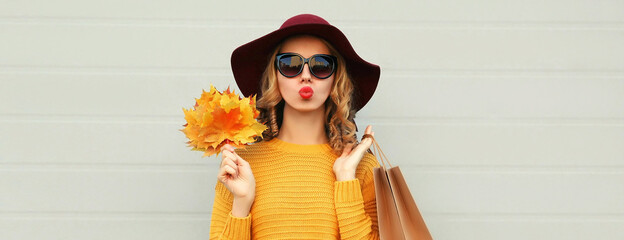 Wall Mural - Autumn portrait of young woman with yellow maple leaves and shopping bags wearing a sweater and hat on gray background