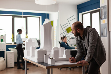 Man working on architectural design at office while looking at laptop computer for industry construction. Project manager inspecting maquette modern design building model