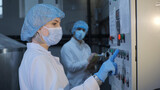Fototapeta Desenie - A woman, an employee of an enterprise or a factory, in a sterile white coat, a medical mask, a disposable cap, hands in disposable latex gloves, presses the equipment button, a colleague in the
