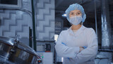 Fototapeta Desenie - A portrait of a woman, an employee of an enterprise or factory, in a sterile white coat, a medical mask, a disposable cap, hands in disposable latex gloves, posing in front of the camera against the