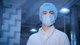 Fototapeta Desenie - Portrait of a young man, a technologist, wearing a sterile mask, disposable cap and a white medical coat in a sterile environment in a production workshop.