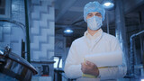 Fototapeta Desenie - In the production workshop, a young technologist with documents in his hands in a sterile medical mask, disposable cap and in a white medical gown is at a large container.