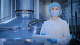 Fototapeta Desenie - Porter of an employee. In a production workshop near a large container, a middle-aged woman, a technologist, with documents in her hands in a sterile medical mask, a disposable cap and a white medical