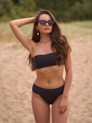 Wall Mural - Attractive young sexy woman wearing black bikini swimwear standing and posing at sandy beach. Stunning girl with wavy brunette hair