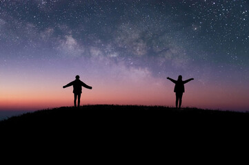 Wall Mural - Silhouette of two tourists standing on top of the mountain and raise both hands watched beautiful night sky, star and milky way. They enjoyed traveling and was successful when he reached the summit.
