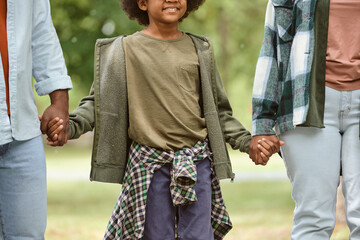 Wall Mural - Smiling African boy holding by hands of his parents while walking in park