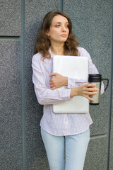 Female portrait of young woman with silver laptop and cup of coffee waiting for a meeting near dark grey wall in the morning, remote job, coffeebreak, smart student concept